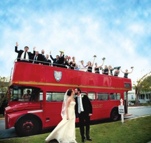 A bride and groom kiss in front of a red double-decker bus with cheering guests on the top deck.