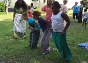 Four children in a sack race outdoors, watched by several adults and kids in the background.