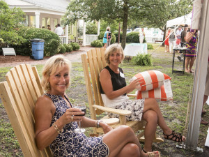 Two women sitting on wooden chairs outdoors, one holding a drink and the other holding a striped pillow.