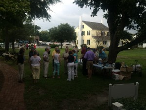 People gather around a table at an outdoor event on grass near a yellow house and trees.