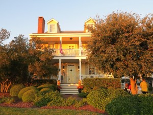 Yellow two-story house with a front porch, American flag, trees, and shrubs in golden sunlight.