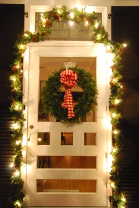 White door decorated with a green wreath with a red bow and glowing garland with pinecones and lights.
