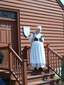 Woman in colonial attire stands on wooden steps outside a brown courthouse building with a sign.