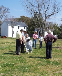 A group of people watch an older woman in historical dress use a wooden tool on a grassy lawn.