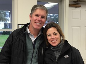 Two people smiling indoors, both wearing jackets, standing closely together near a door and window.