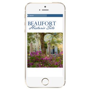 A smartphone displays the Beaufort Historic Site website with flowers and trees in the background.
