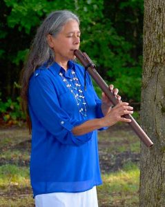 Woman in a blue shirt plays a wooden flute outdoors near a tree, with greenery in the background.