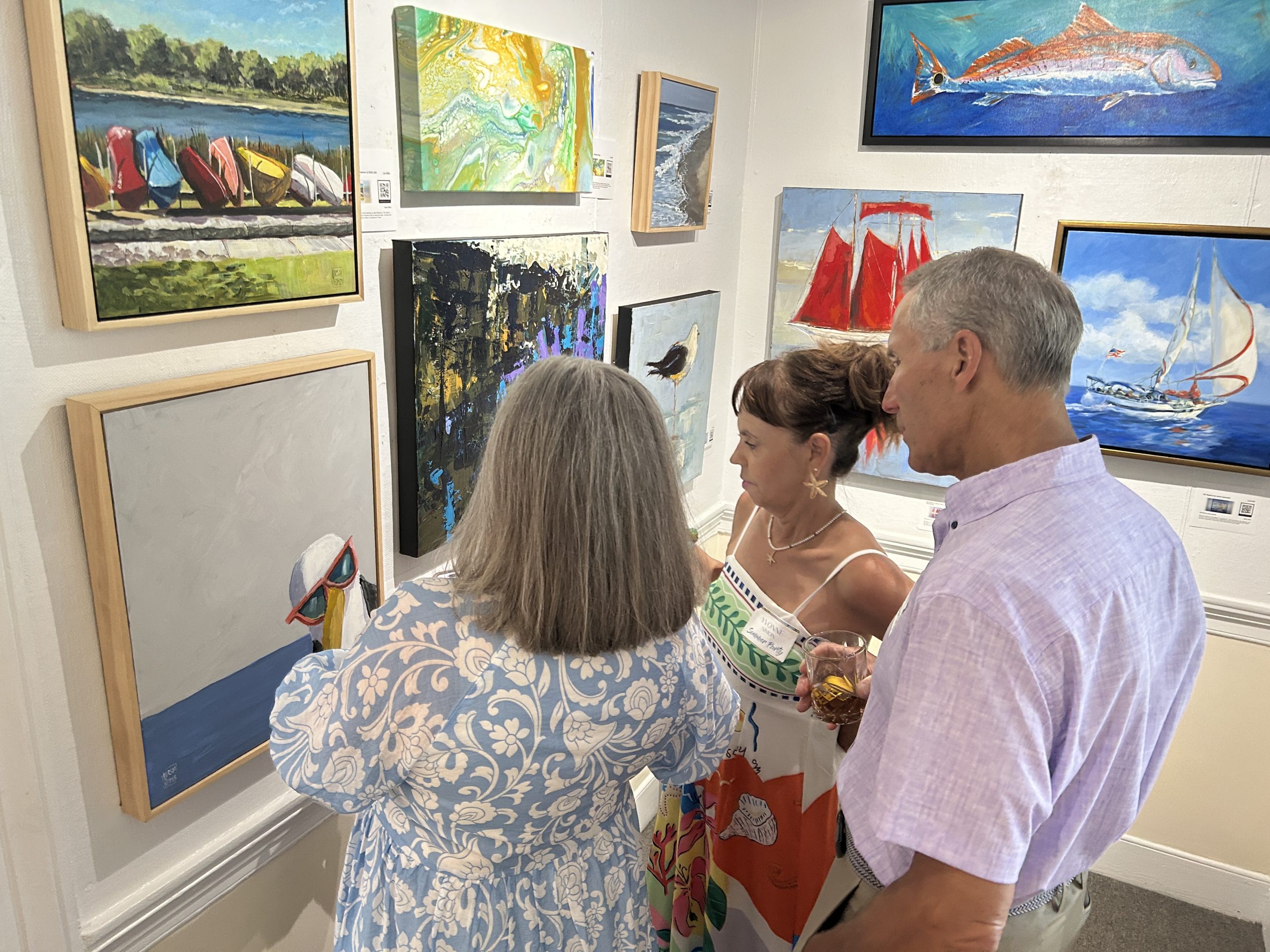 Three people look at paintings displayed on a gallery wall.