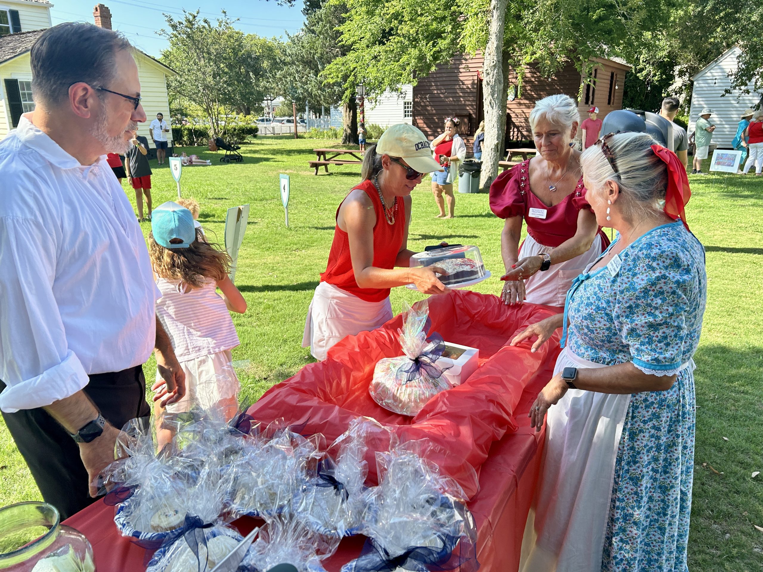 People at an outdoor table setting out items on a red tablecloth.